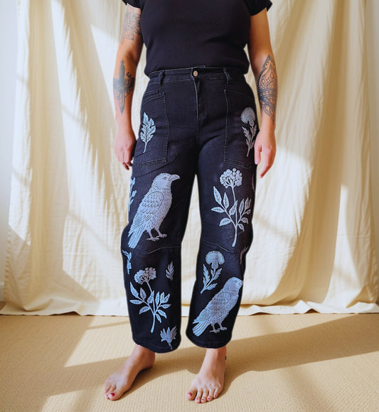 A person stands barefoot in front of a cream fabric backdrop wearing dark jeans with large white bird and floral embroidery, paired with a black short-sleeve top.