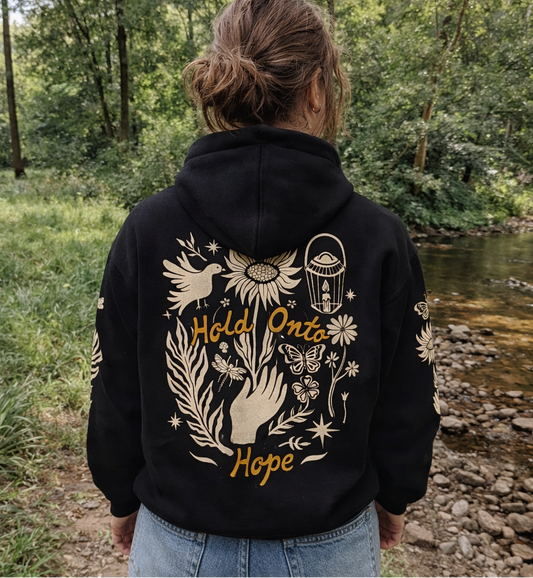 Person wearing a black hoodie with gold folk nature designs and 'Hold Onto Hope' text, standing in a natural setting.
