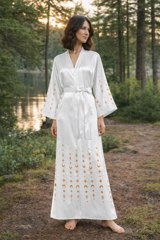 Woman in a white kimono robe with gold ornamental patterns standing in a forest by a lake.