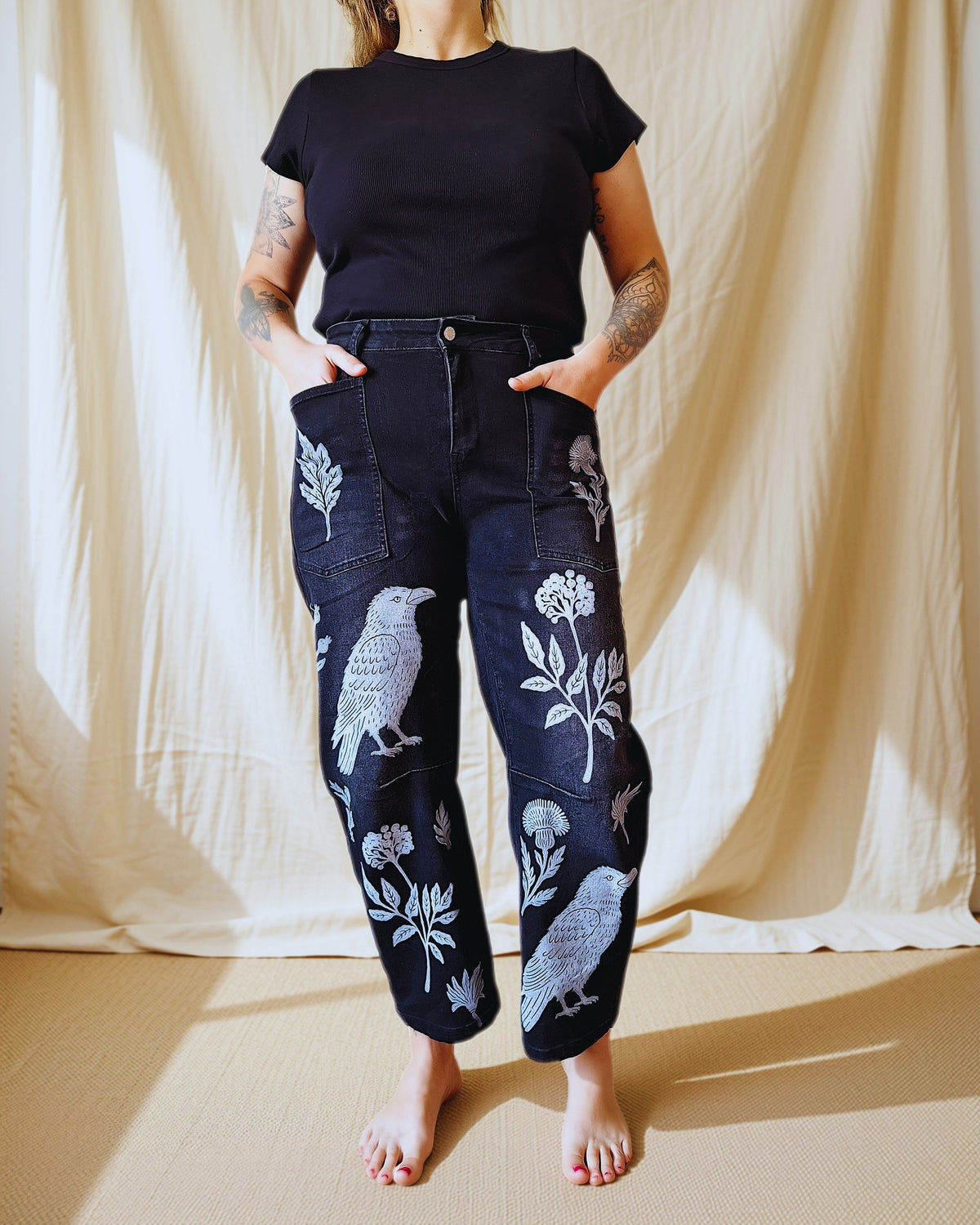 A model stands barefoot in front of a draped beige fabric backdrop, wearing dark indigo jeans with white bird and floral embroidery, paired with a black t-shirt.