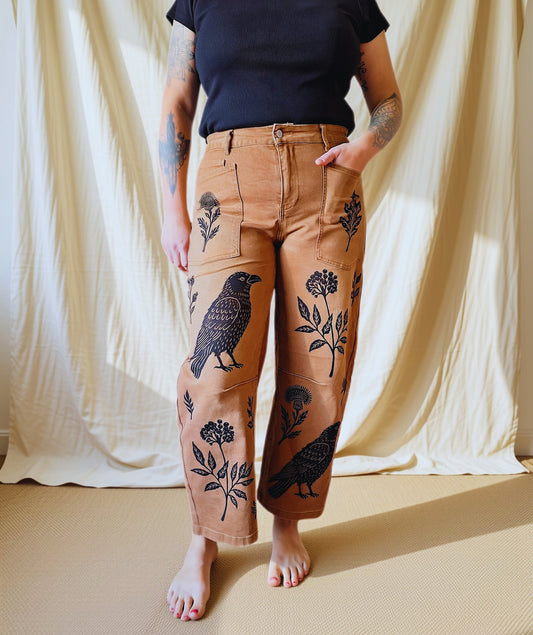 A person stands barefoot in front of a draped cream fabric backdrop, wearing tan wide-leg pants with black bird and floral illustrations, paired with a dark navy short-sleeve top.