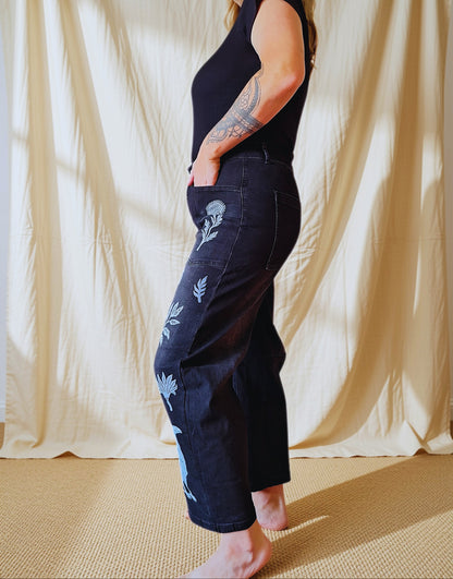 A woman stands barefoot in profile wearing dark indigo denim cropped pants with light blue floral embroidery down the side, paired with a black sleeveless top, set against a draped cream fabric backdrop.