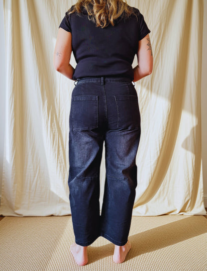 A person standing with their back to the camera, wearing dark indigo wide-leg denim jeans and a black t-shirt, posed in front of a draped cream fabric backdrop.
