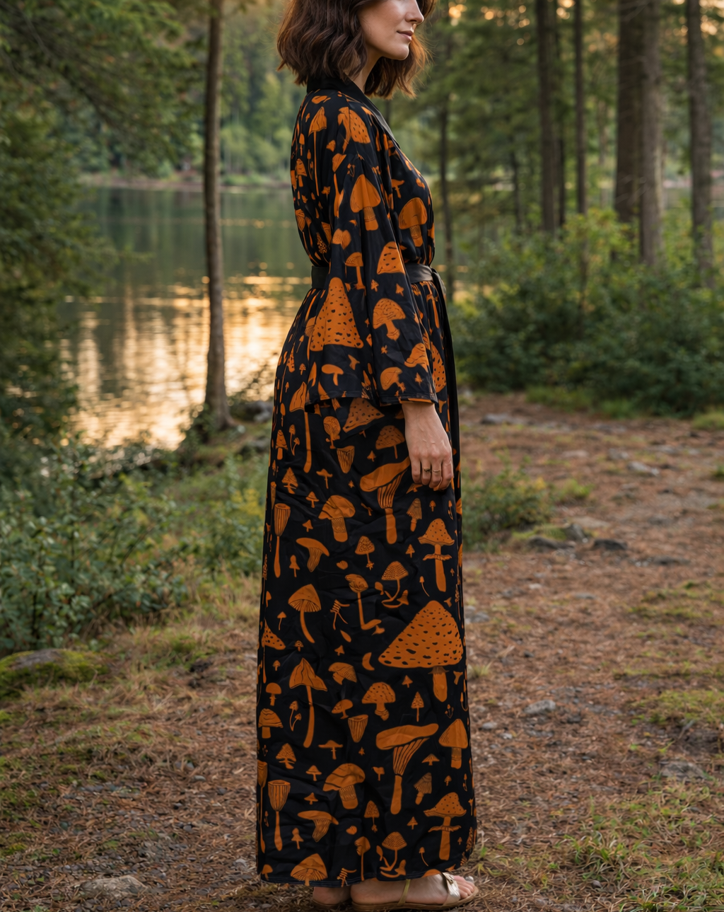 Woman in a long kimono dress with mushroom pattern standing in a forest by a lake.