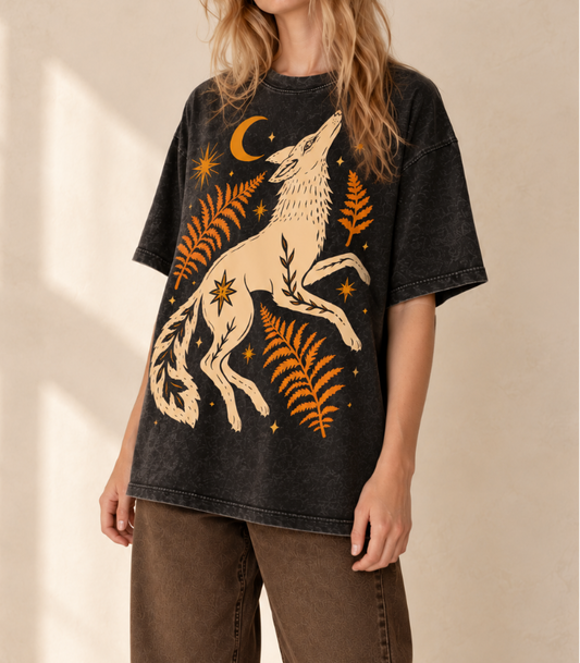 A woman wearing a faded black graphic t-shirt with a cream and orange print of a howling wolf, ferns, and stars, paired with brown pants against a warm beige wall.