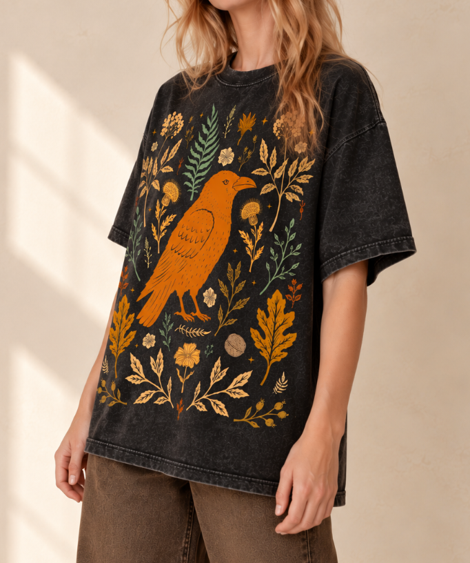 A woman wearing a faded black graphic t-shirt featuring an orange bird and yellow, green, and beige botanical illustration, paired with brown wide-leg pants against a warm beige wall.