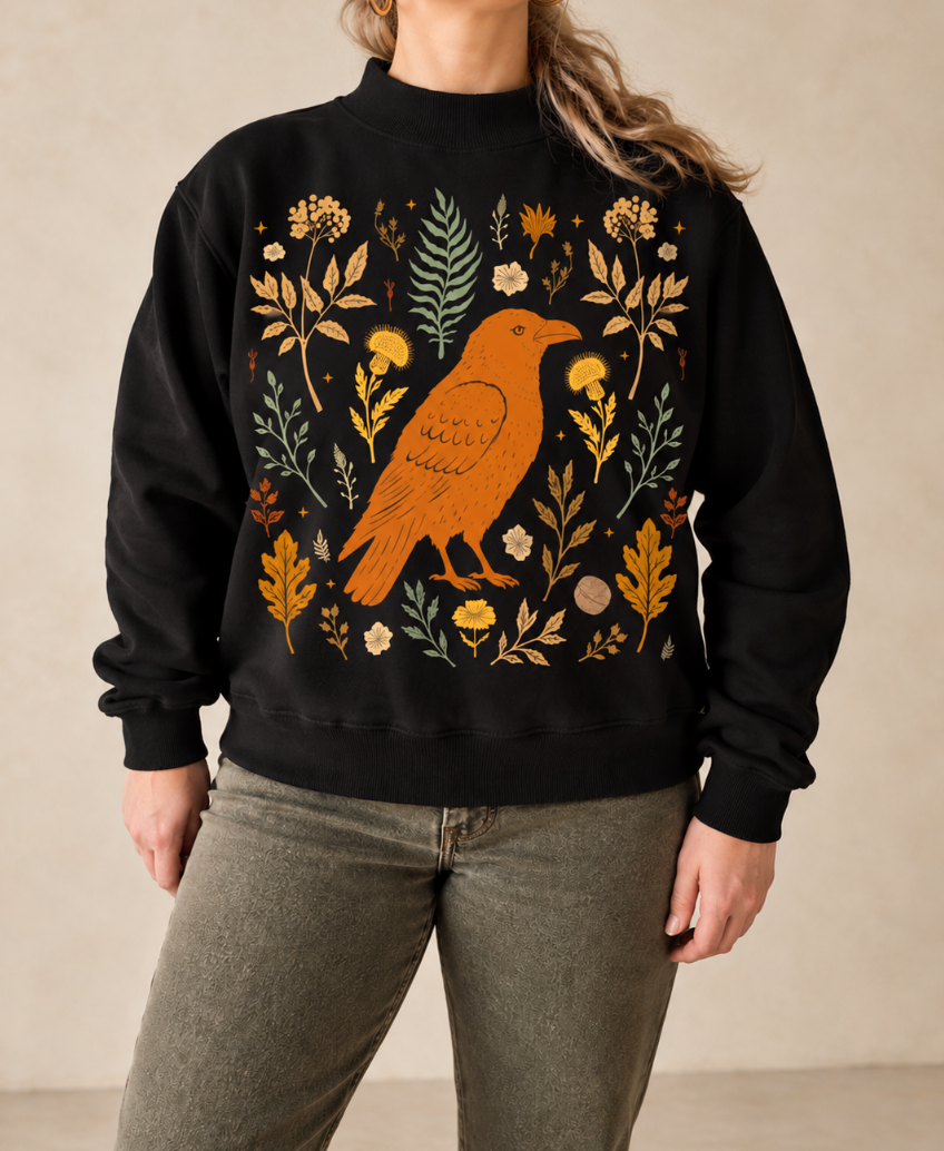 A person wearing a black crewneck sweatshirt with a large orange bird and autumn floral pattern on the front, paired with olive green pants, set against a neutral beige background.