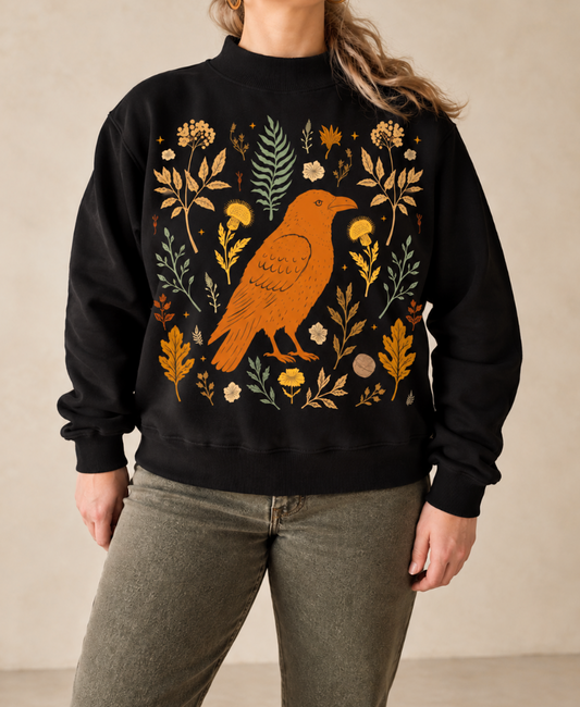 A person wearing a black crewneck sweatshirt with a large orange bird and autumn floral pattern on the front, paired with olive green pants, set against a neutral beige background.