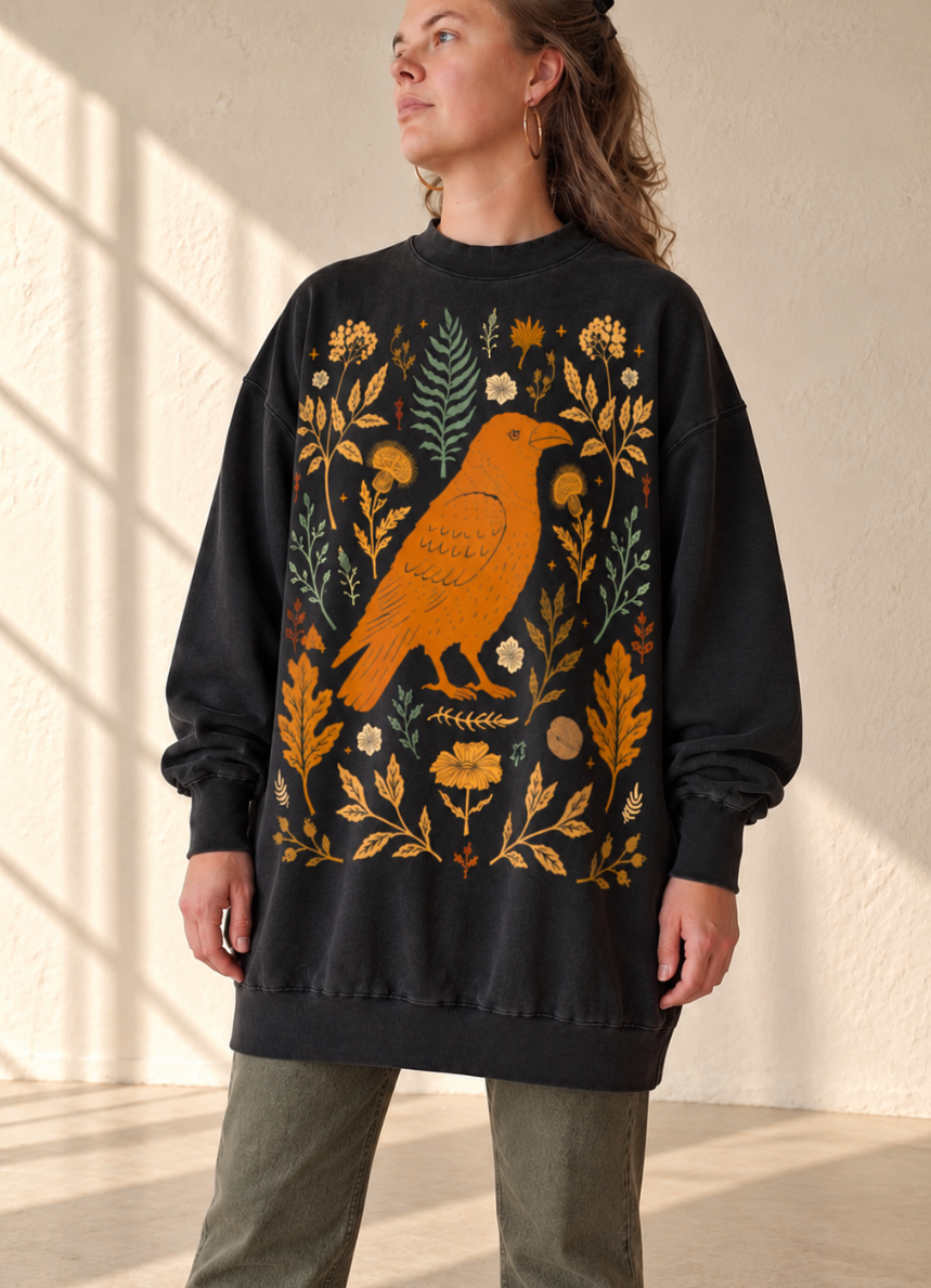 A woman wearing a black crewneck sweatshirt with a large graphic print of an orange bird surrounded by yellow, green, and white botanical elements. She is standing in a sunlit room with a textured beige wall.