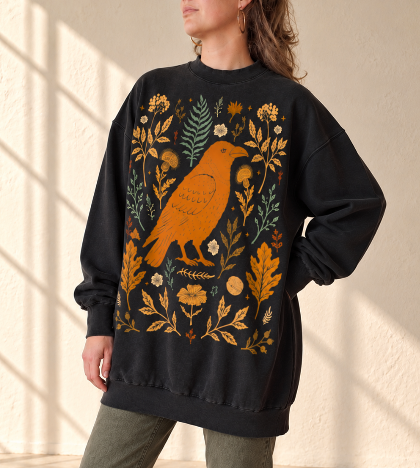 A woman wearing an oversized black sweatshirt with a large front graphic of an orange bird surrounded by yellow, beige, and muted green botanical details, photographed indoors against a textured cream wall with soft window shadows.