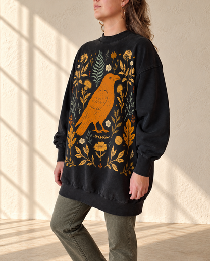 A woman wearing a black crewneck sweatshirt with a large orange bird and floral botanical pattern on the front, paired with olive green pants, standing in a sunlit room with textured walls.