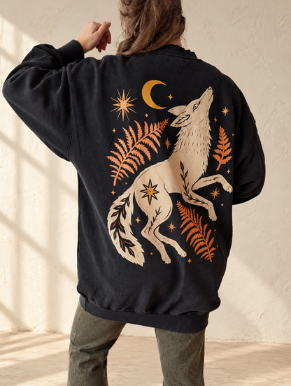 A person shown from the back wearing a black oversized bomber-style jacket with a large cream and orange graphic of a howling wolf, ferns, stars, and a crescent moon, standing against a textured off-white wall.