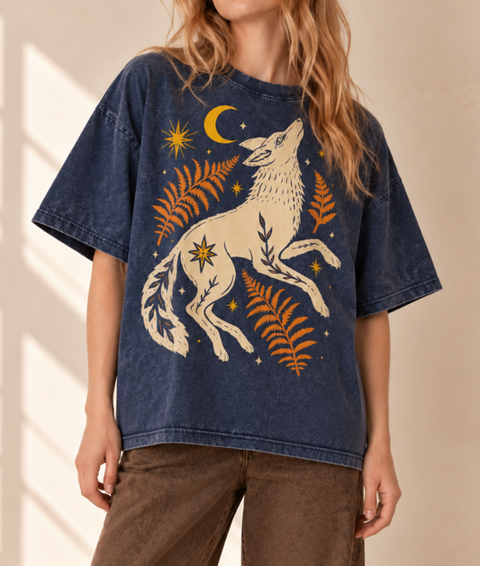 A woman wearing a dark blue washed graphic t-shirt featuring a cream-colored howling wolf surrounded by orange ferns, yellow stars, and a crescent moon, paired with brown pants.