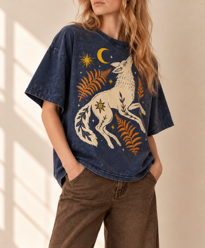 A woman wearing a faded dark blue graphic t-shirt with a cream-colored wolf, yellow crescent moon, and orange ferns, paired with brown corduroy pants, set against a warm beige background with soft window shadows.
