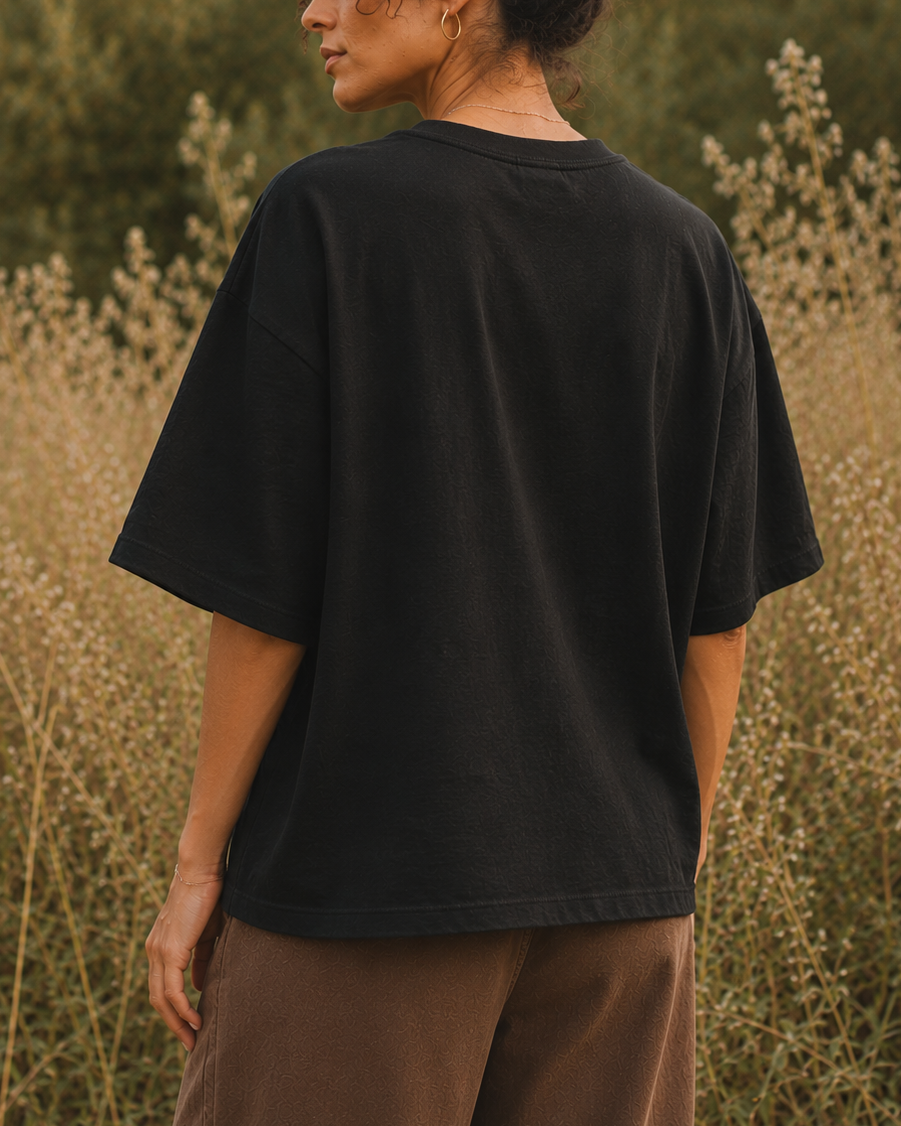 A woman wearing an oversized black t-shirt and brown pants, photographed from behind in a field of dry grass with trees in the background.