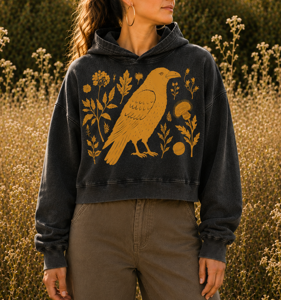 A woman wearing a dark charcoal gray hooded sweatshirt with a large mustard yellow bird and floral graphic, paired with brown wide-leg pants, standing in a field of dry plants at sunset.
