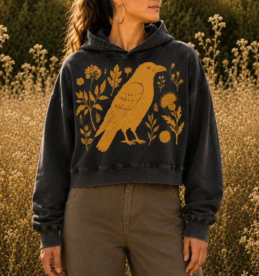 A woman wearing a dark charcoal gray hooded sweatshirt with a large mustard yellow bird and floral graphic, paired with brown wide-leg pants, standing in a field of dry plants at sunset.
