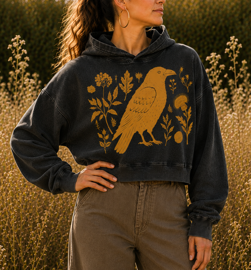 A woman wearing a washed black cropped hoodie with a gold raven and floral graphic, paired with brown wide-leg pants, standing in a field of golden wildflowers at sunset.