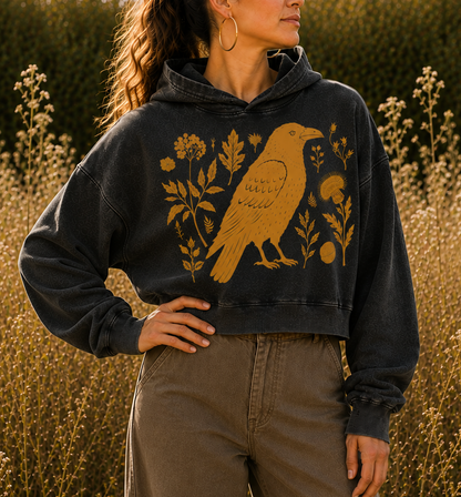 A woman wearing a washed black cropped hoodie with a gold raven and floral graphic, paired with brown wide-leg pants, standing in a field of golden wildflowers at sunset.