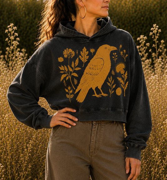 A woman wearing a washed black cropped hoodie with a gold raven and floral graphic, paired with brown wide-leg pants, standing in a field of golden wildflowers at sunset.