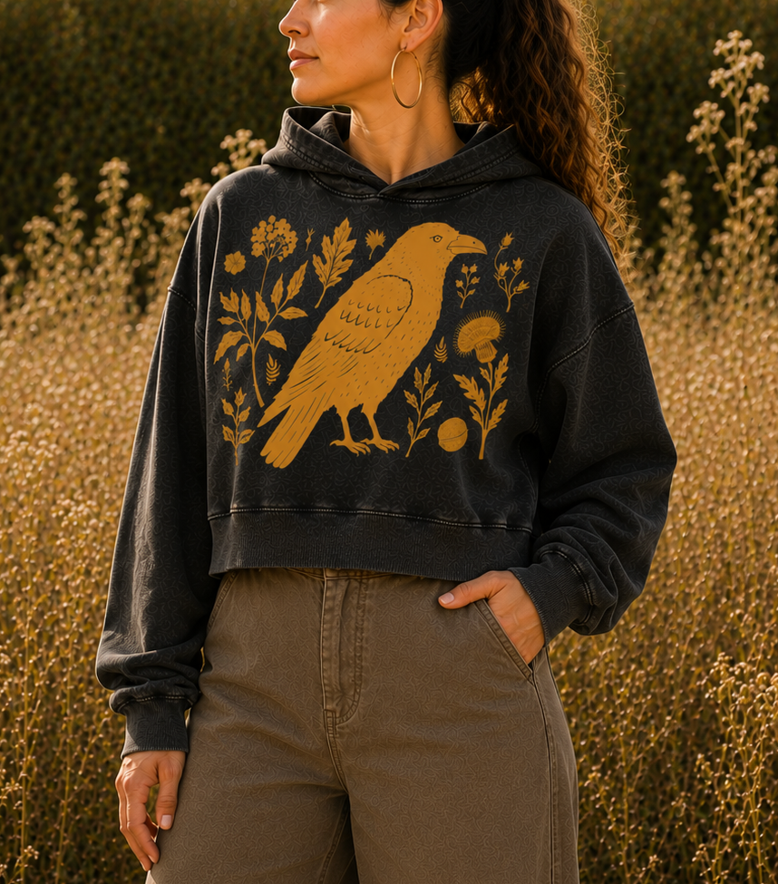 A woman wearing a dark charcoal gray cropped hoodie with a yellow bird and floral graphic, paired with brown wide-leg pants, standing in a field of dry grass.