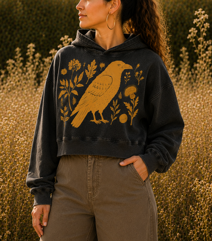 A woman wearing a dark charcoal gray cropped hoodie with a yellow bird and floral graphic, paired with brown wide-leg pants, standing in a field of dry grass.