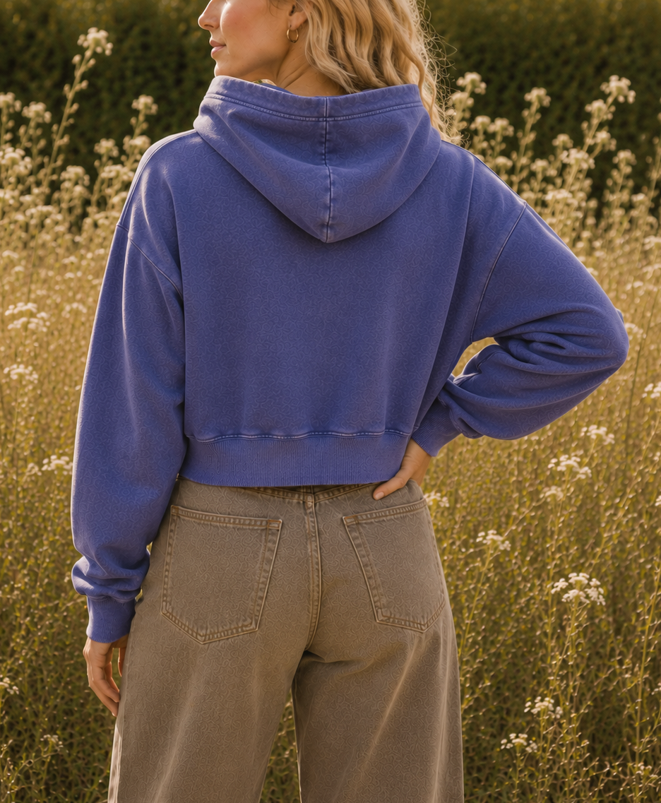 A woman standing in a sunlit field of wildflowers, photographed from the back and wearing a cropped blue-purple hooded sweatshirt and light brown pants.