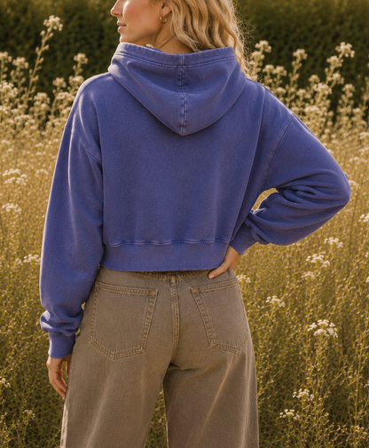 A woman standing in a sunlit field of wildflowers, photographed from the back and wearing a cropped blue-purple hooded sweatshirt and light brown pants.