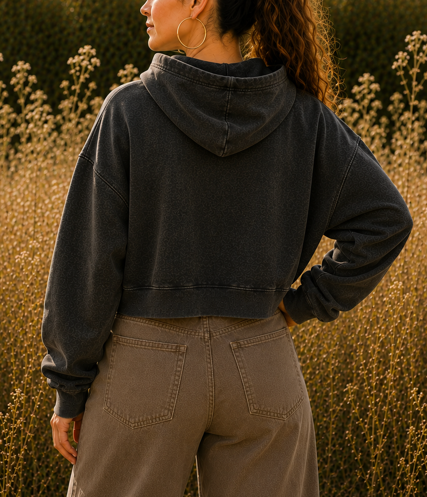A woman stands in a field of dry grass, photographed from the back, wearing a faded charcoal gray cropped hoodie and beige wide-leg pants.