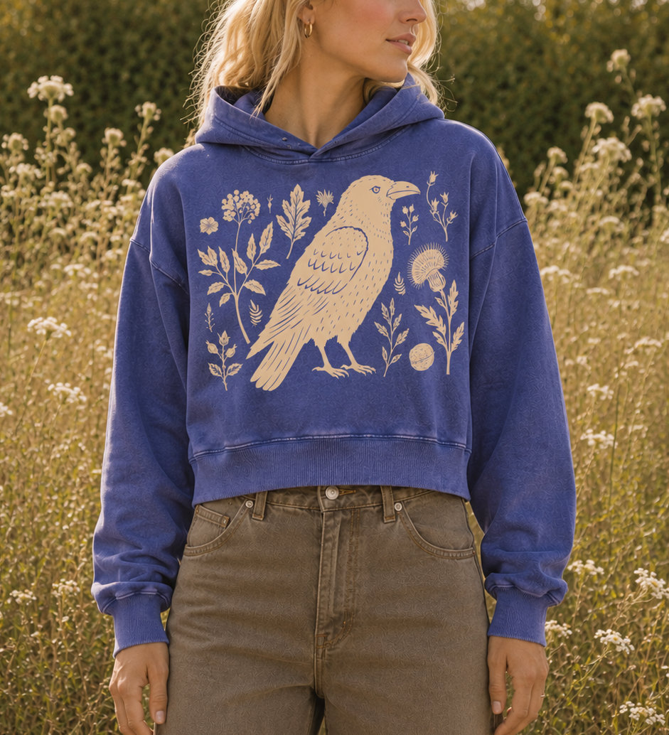 A woman wearing a cropped blue hoodie with a large cream bird and floral graphic, standing in a field of wildflowers outdoors.