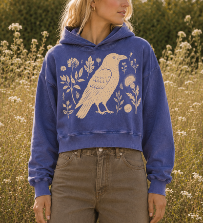 A woman wearing a cropped blue hoodie with a large cream bird and floral graphic, standing in a field of wildflowers outdoors.