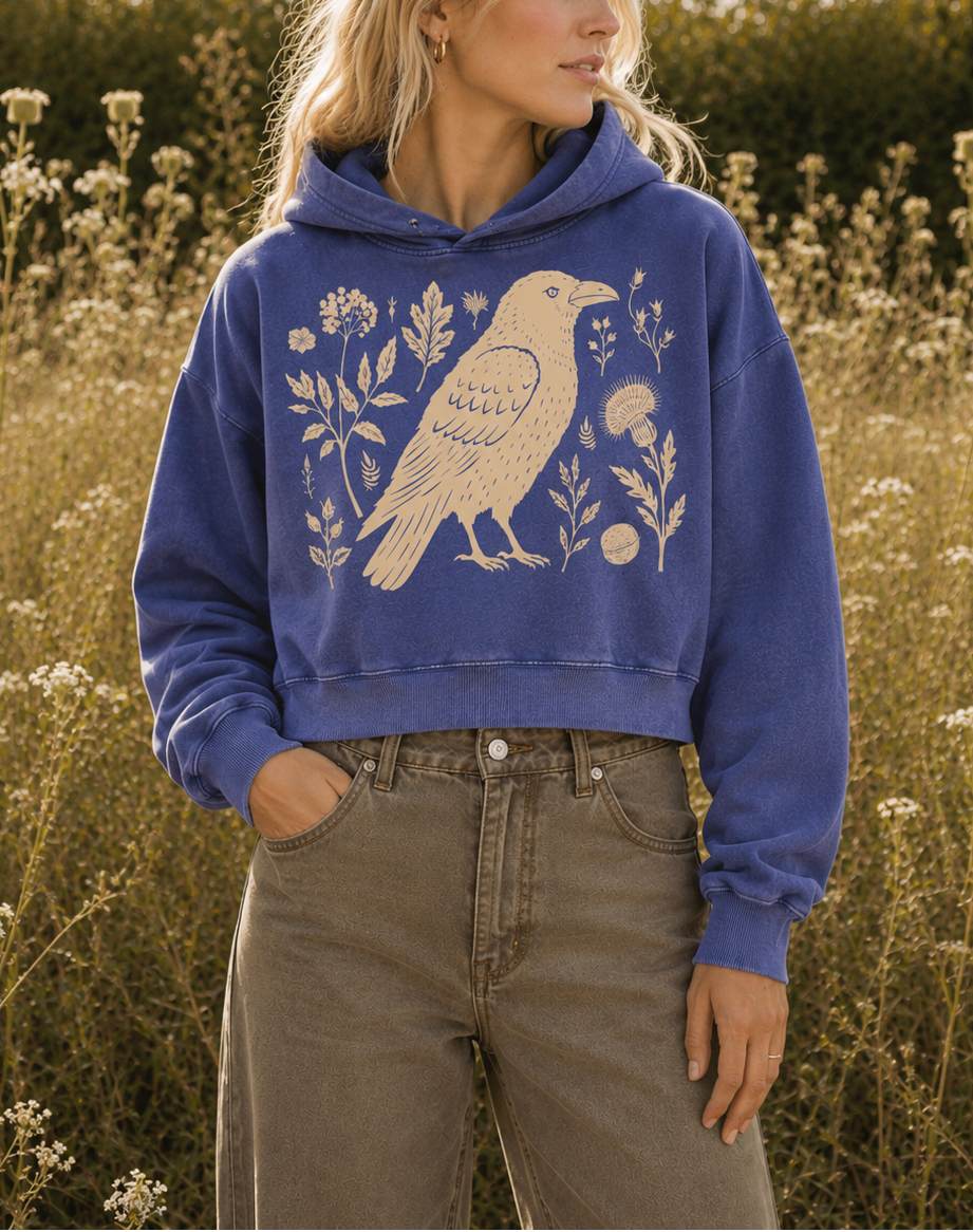 A woman wearing a cropped blue hoodie with a cream-colored bird and floral graphic, standing in a field of wildflowers.