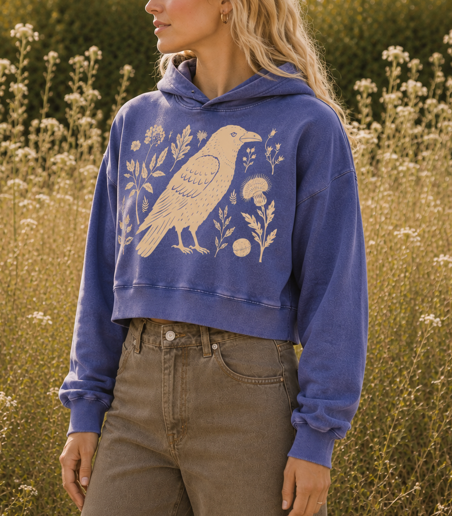 A woman standing in a sunlit field of wildflowers, wearing a cropped blue hoodie with a large cream bird and floral print on the front, paired with brown pants.