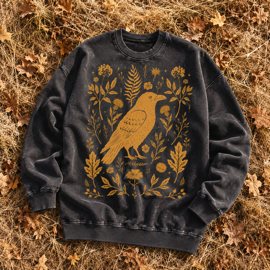 A dark washed crewneck sweatshirt featuring a golden-yellow raven surrounded by botanical illustrations, laid flat on a bed of dry autumn leaves.