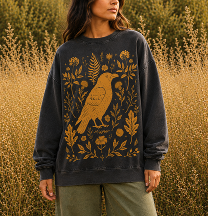 A woman wearing a dark charcoal gray sweatshirt with a large golden-brown raven and botanical graphic on the front, standing in a field of dry golden grass.