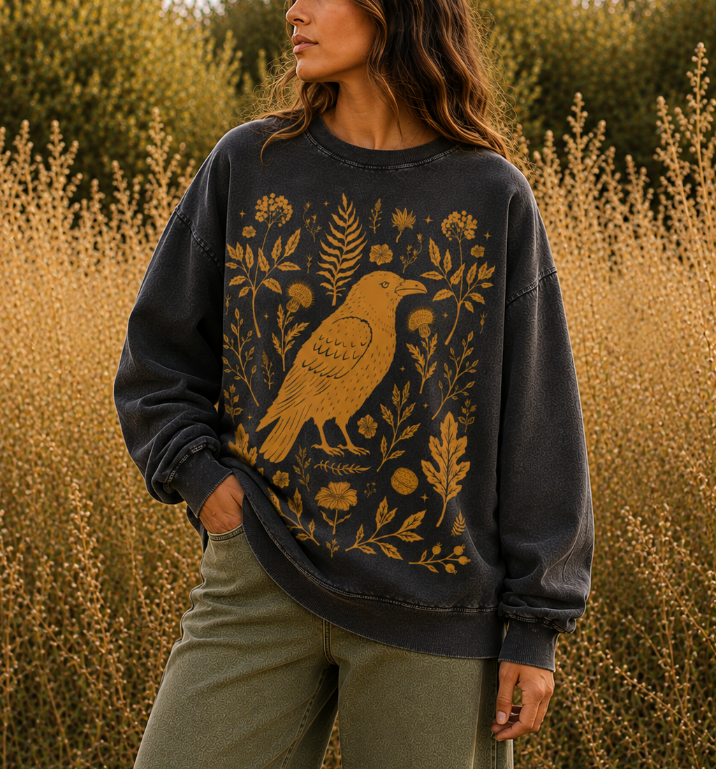 A woman wearing a dark charcoal gray crewneck sweatshirt with a large mustard yellow bird and botanical print, standing in a field of dry golden plants.