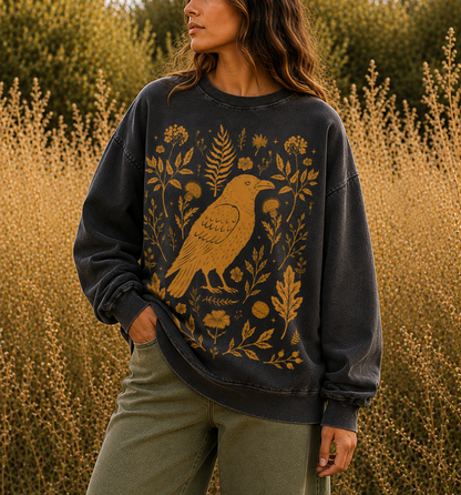 A woman wearing a dark charcoal gray crewneck sweatshirt with a large mustard yellow bird and botanical print, standing in a field of dry golden plants.