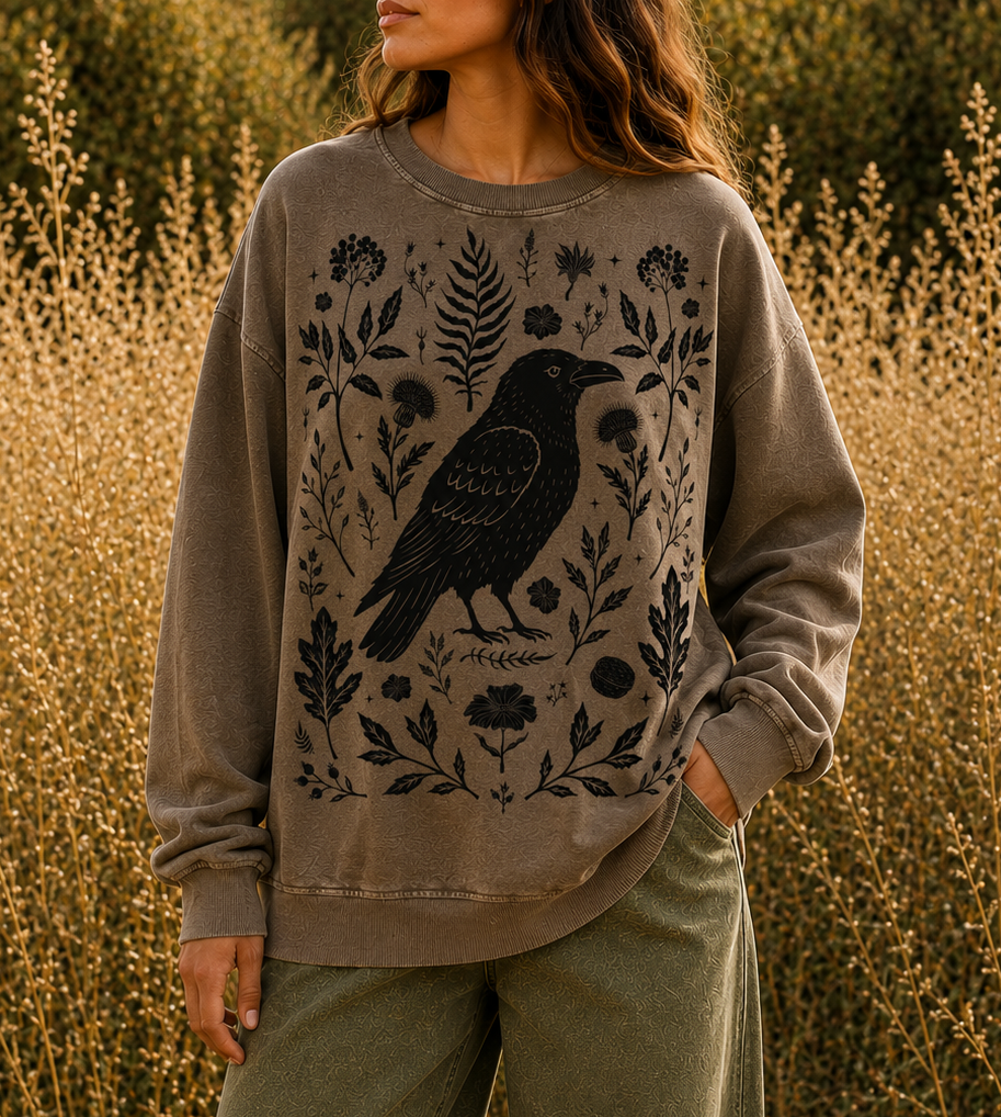 A woman wearing a taupe crewneck sweatshirt with a black raven and botanical graphic, standing in a field of dry golden grass.