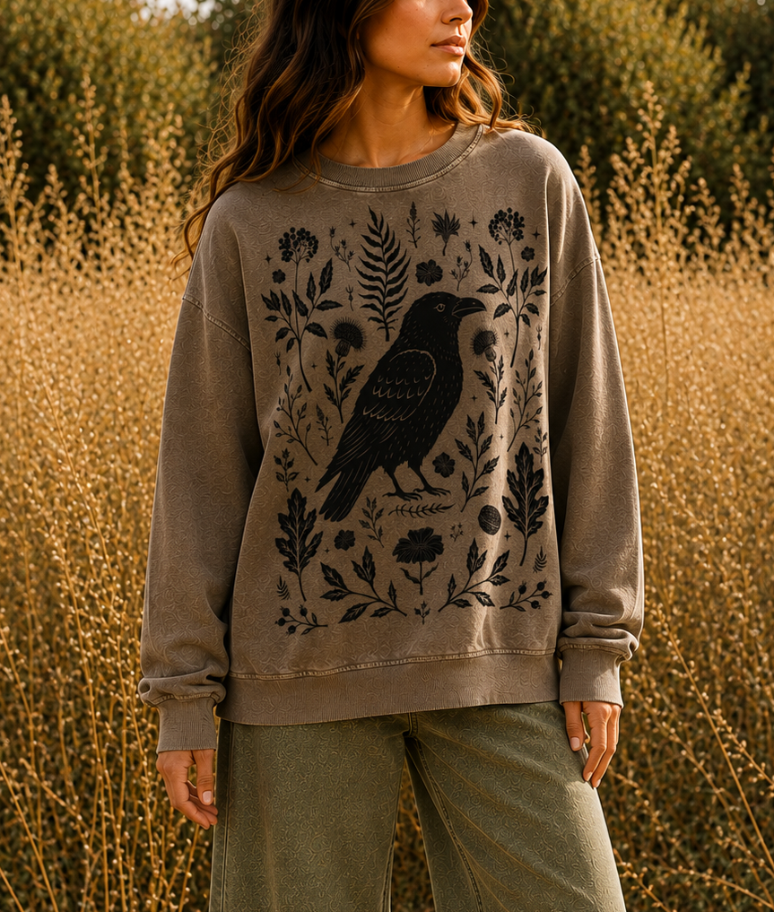 A woman stands in a field of dry golden plants wearing a taupe crewneck sweatshirt with a black raven and botanical graphic, paired with olive green pants.