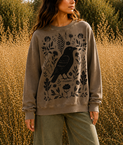 A woman stands in a field of dry golden plants wearing a taupe crewneck sweatshirt with a black raven and botanical graphic, paired with olive green pants.