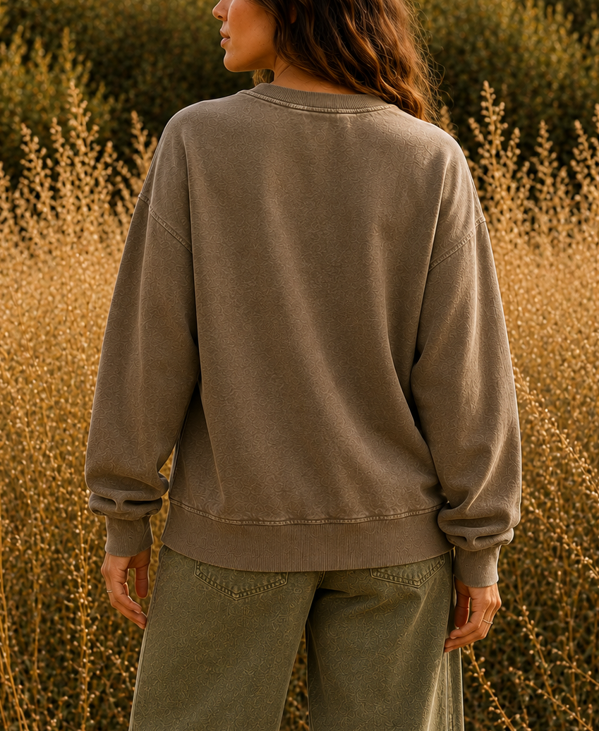 A woman stands outdoors in a field of dry golden grass, photographed from behind, wearing a muted taupe-brown crewneck sweatshirt and olive green pants.