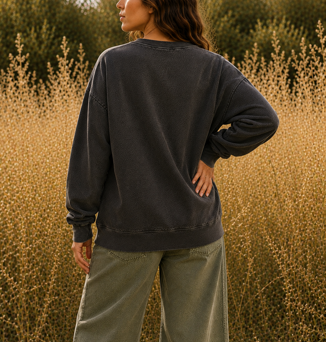 A woman stands outdoors in a field of dry golden grass, photographed from the back. She is wearing a dark washed charcoal sweatshirt and olive green wide-leg pants.