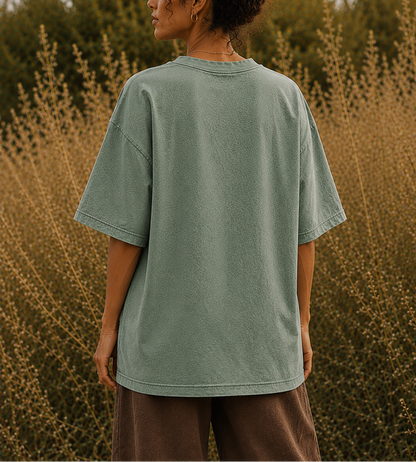 A woman stands outdoors in a field of dry grass, viewed from behind, wearing an oversized sage green t-shirt and brown pants.