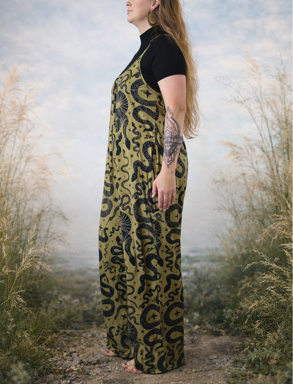 Woman wearing a long, patterned snake jumpsuit standing in a natural setting with tall grass and a cloudy sky.
