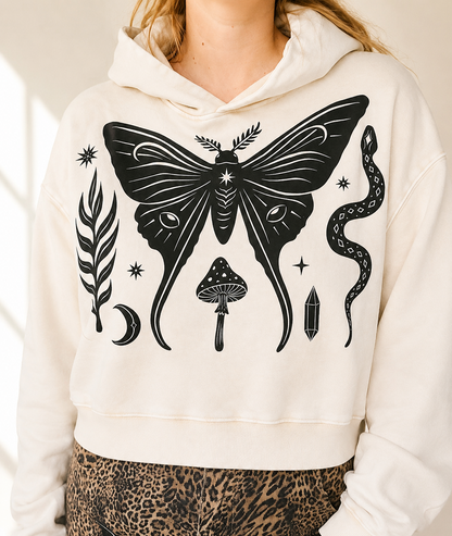 A person wearing a cream-colored cropped hoodie with a black graphic print of a butterfly, mushroom, snake, crystal, and moon, paired with leopard-print pants.