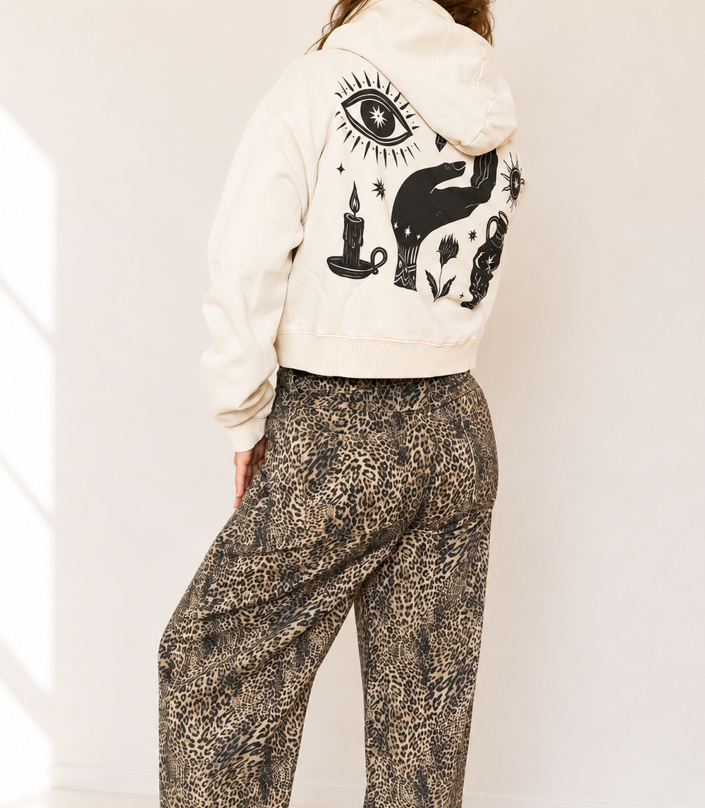 A model shown from the back wearing a cream-colored cropped hoodie with a black occult-style graphic print on the back, paired with brown and beige leopard-print wide-leg pants.
