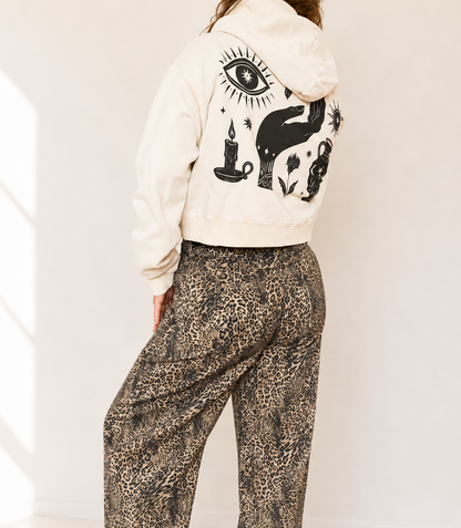 A model shown from the back wearing a cream-colored cropped hoodie with a black occult-style graphic print on the back, paired with brown and beige leopard-print wide-leg pants.