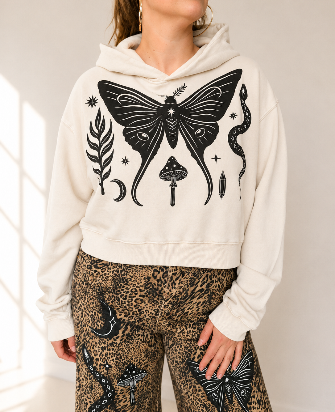 A woman wearing a cream-colored cropped hoodie with a large black graphic print of a moth, mushroom, snake, and moon on the front, paired with leopard-print pants.