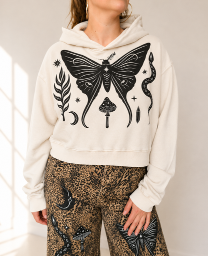A woman wearing a cream-colored cropped hoodie with a large black graphic print of a moth, mushroom, snake, and moon on the front, paired with leopard-print pants.
