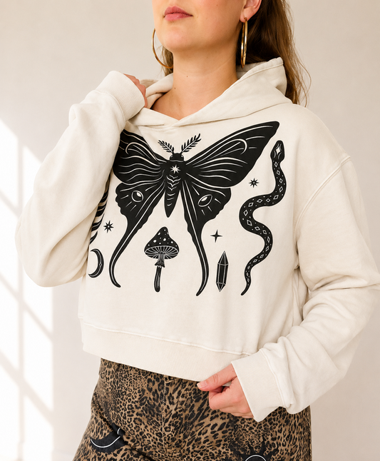A woman wearing a cream-colored cropped hoodie with a large black gothic-style graphic of a moth, snake, and mushroom on the front, paired with leopard-print pants.